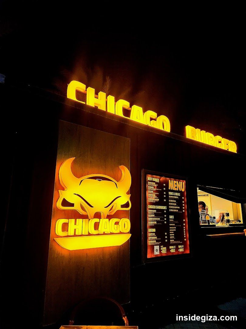 A brightly lit storefront featuring a glowing yellow neon sign with the word 'CHICAGO' and a bull logo. Below, there is a menu board displaying various food items like burgers and fries. The scene captures the vibrant nighttime ambiance of a casual dining establishment.