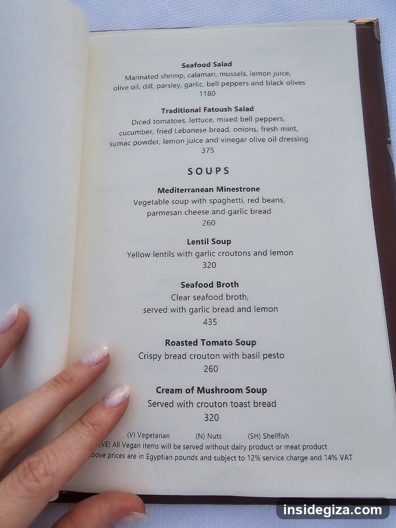 A close-up photo of an open menu book showing a section titled 'Soups'. A hand with light pink nail polish is visible on the left side, holding or resting near the page.