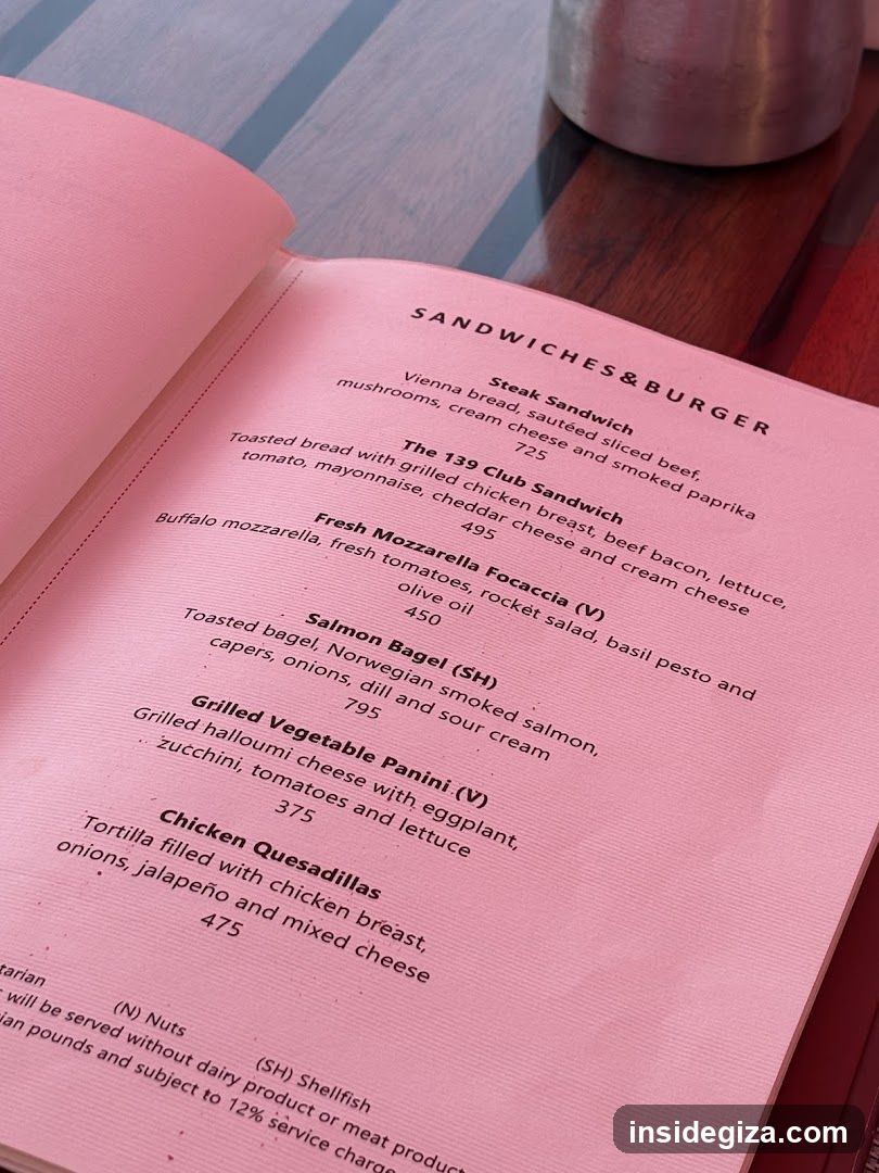 A close-up photo of an open pink menu book on a wooden table, showing the 'SANDWICHES & BURGER' section with items like Steak Sandwich (725), The 139 Club Sandwich (495), Fresh Mozzarella Focaccia (V) (450), Salmon Bagel (SH) (795), Grilled Vegetable Panini (V) (375), and Chicken Quesadillas (475). A metal cup is visible in the background.