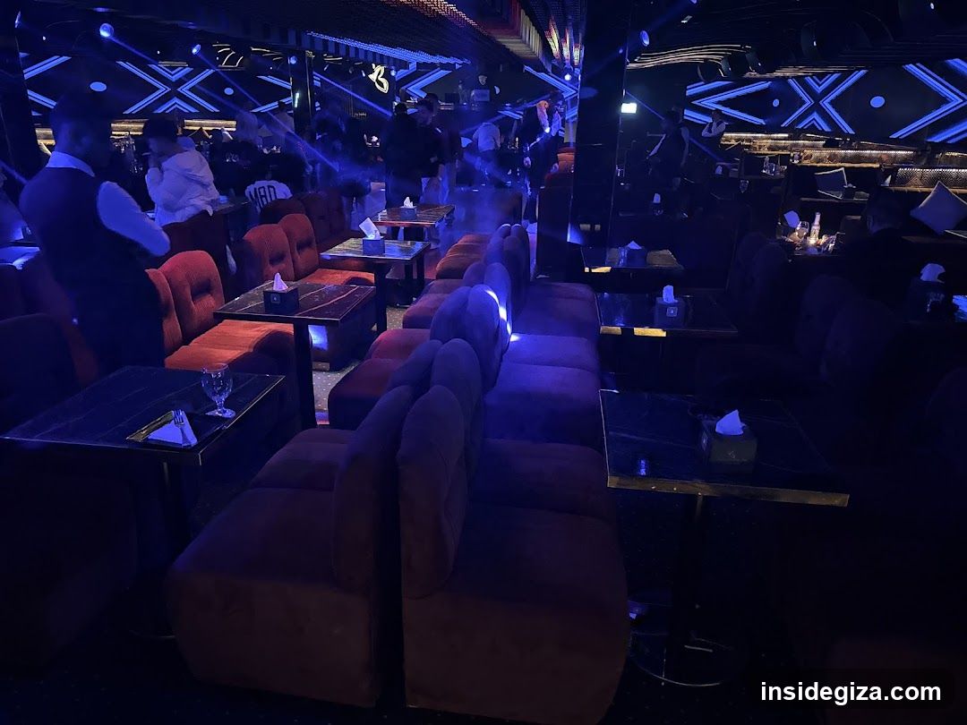 The image shows the interior of a dimly lit club or lounge, featuring rows of plush red and purple sofas arranged around small tables. The space is illuminated by blue LED lights on the ceiling, creating a vibrant atmosphere. A staff member in a vest stands near the entrance, and patrons are seated at various tables. There are no visible menus or shop fronts.