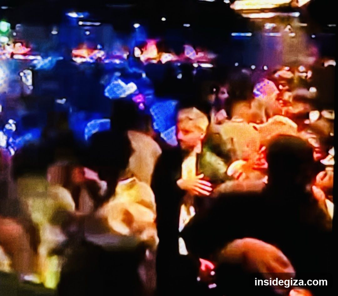 A blurry, low-resolution image of a crowded indoor venue at night. The scene is illuminated by colorful stage lights—primarily blue and orange/red hues. A central figure with blond hair appears to be wearing formal attire (possibly a tuxedo or suit). The crowd is dense, with many people visible in the background, some holding drinks or phones. No signage, menus, or shop fronts are identifiable.