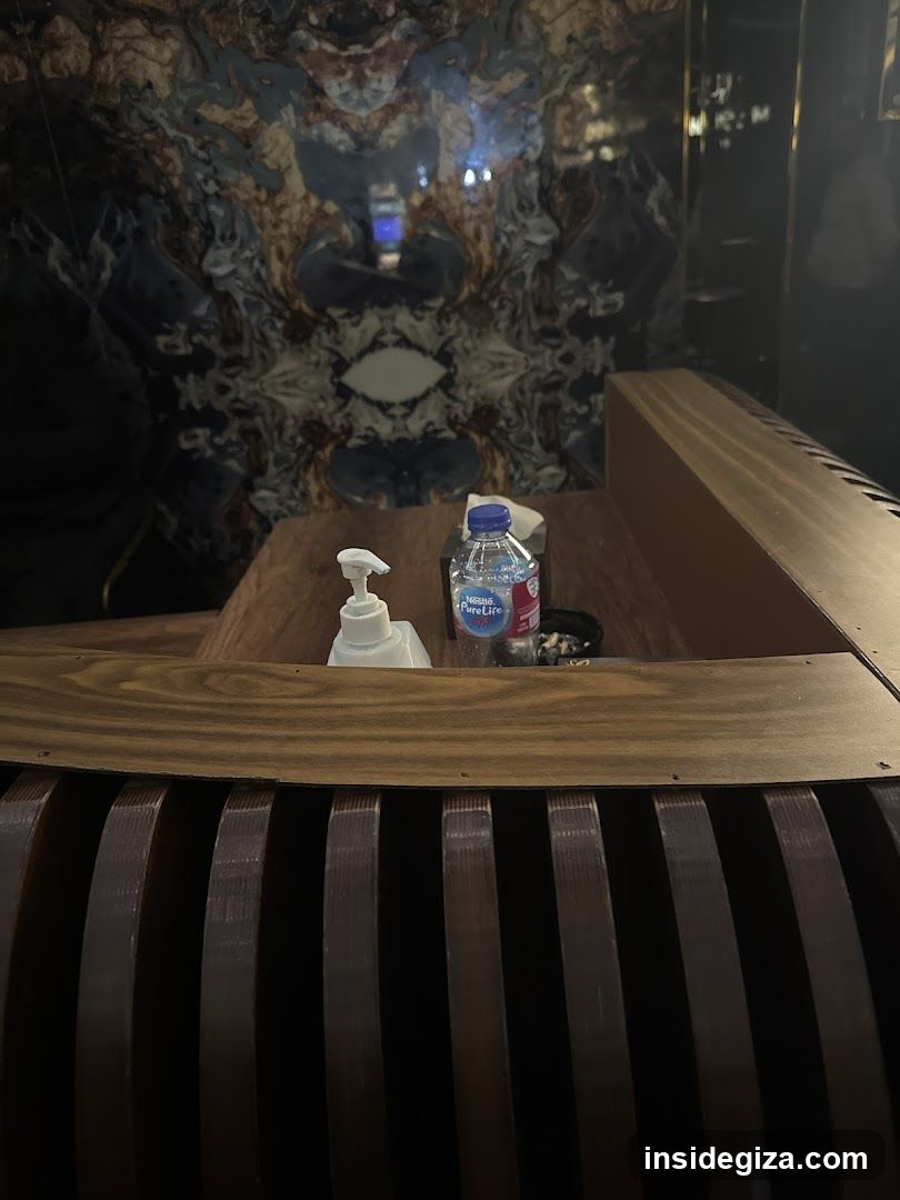 A wooden table in a dimly lit interior space, featuring a pump bottle of hand sanitizer and a plastic water bottle labeled 'Pure Life'. The background includes an ornate, patterned wall or artwork. A slatted wooden structure is visible in the foreground.