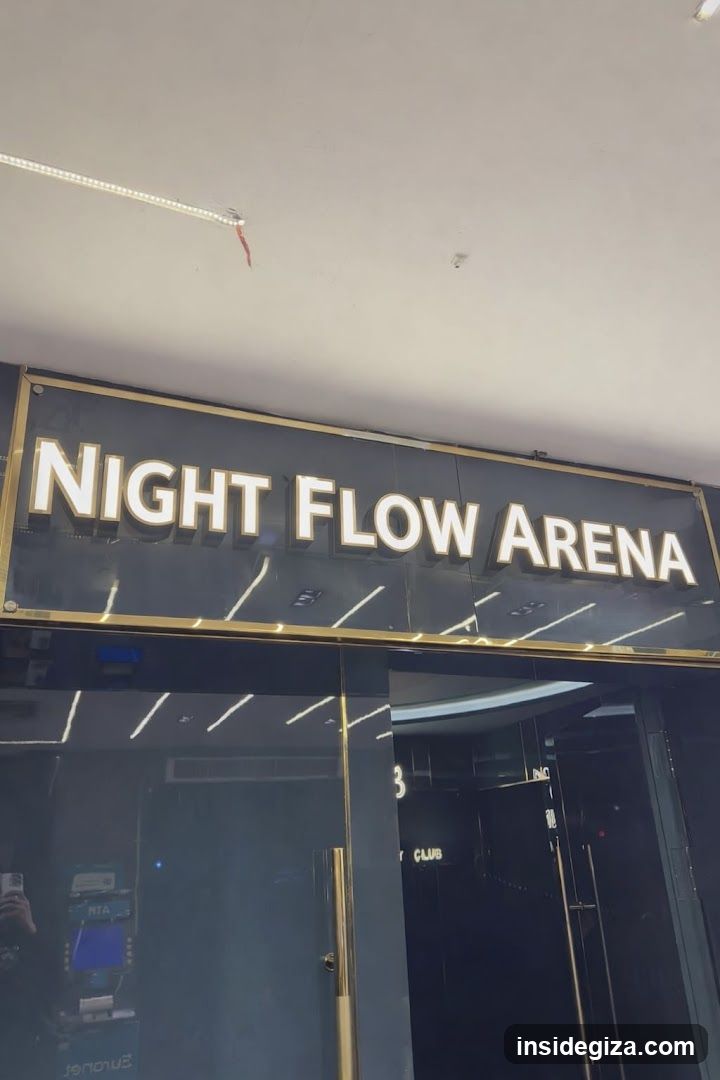 Exterior shot of a storefront with illuminated signage reading 'NIGHT FLOW ARENA' in bold white letters on a dark background framed by gold trim. Glass doors and windows reflect interior lighting, including linear ceiling lights. A partial view of an ATM or kiosk is visible through the left window.