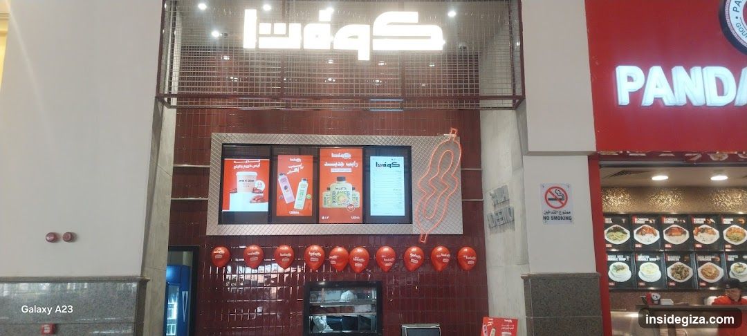 A shop front featuring a large illuminated sign in Arabic script above the entrance. Below, there are four digital screens displaying advertisements or menus. A row of red balloons is strung across the lower part of the facade. To the right, another establishment named 'PANDA' is visible. The wall behind the screens has a textured pattern.