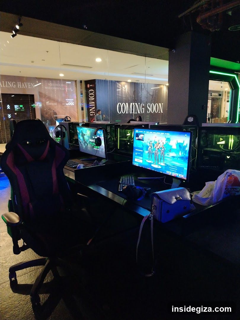 Interior of a gaming lounge with multiple computer setups, each featuring large monitors displaying games and keyboards. A prominent racing-style gaming chair is positioned in the foreground. In the background, signage reads 'COMING SOON' on a wall, suggesting an upcoming event or opening. The environment includes ambient lighting and promotional posters visible through glass partitions.