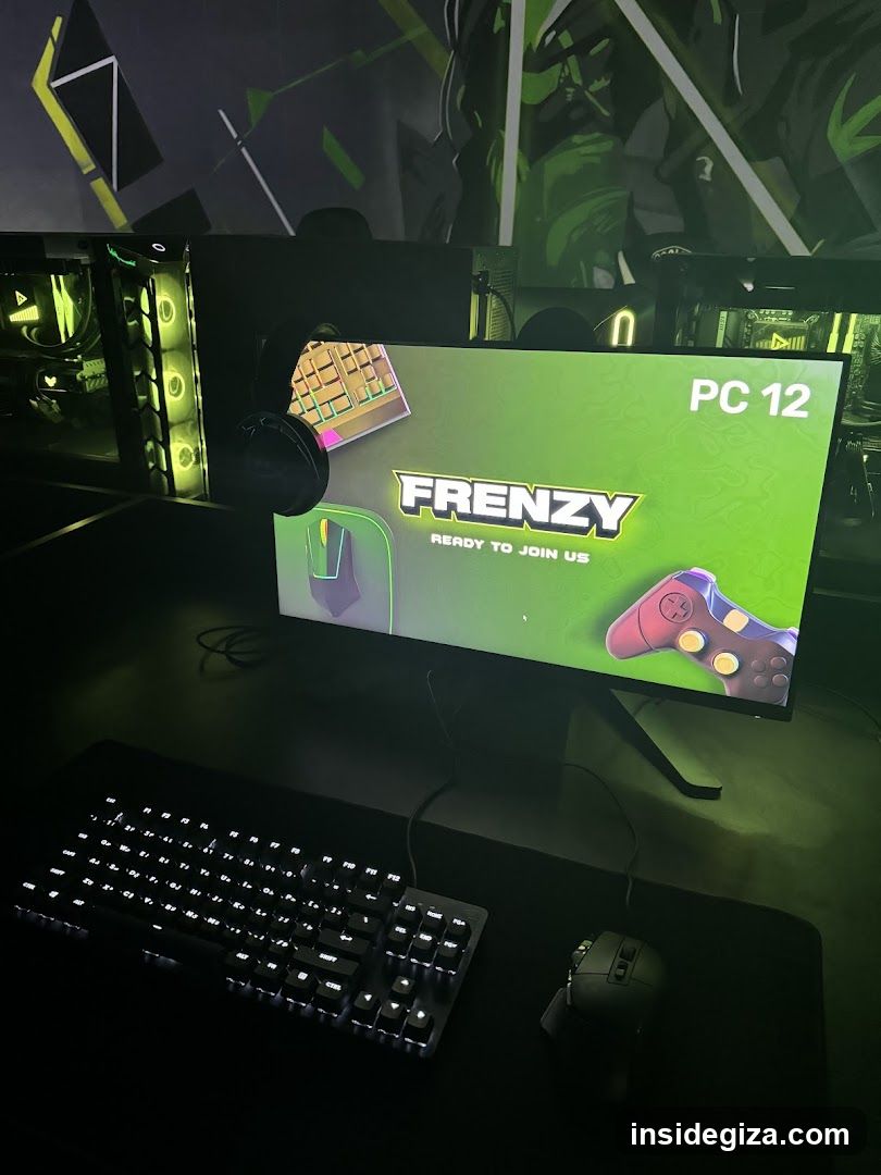 A close-up view of a gaming setup featuring a monitor displaying the word 'FRENZY' with a green background and images of a keyboard, mouse, and game controller. The desk includes a backlit mechanical keyboard and a gaming mouse. In the background, there are computer towers with glowing green lighting.