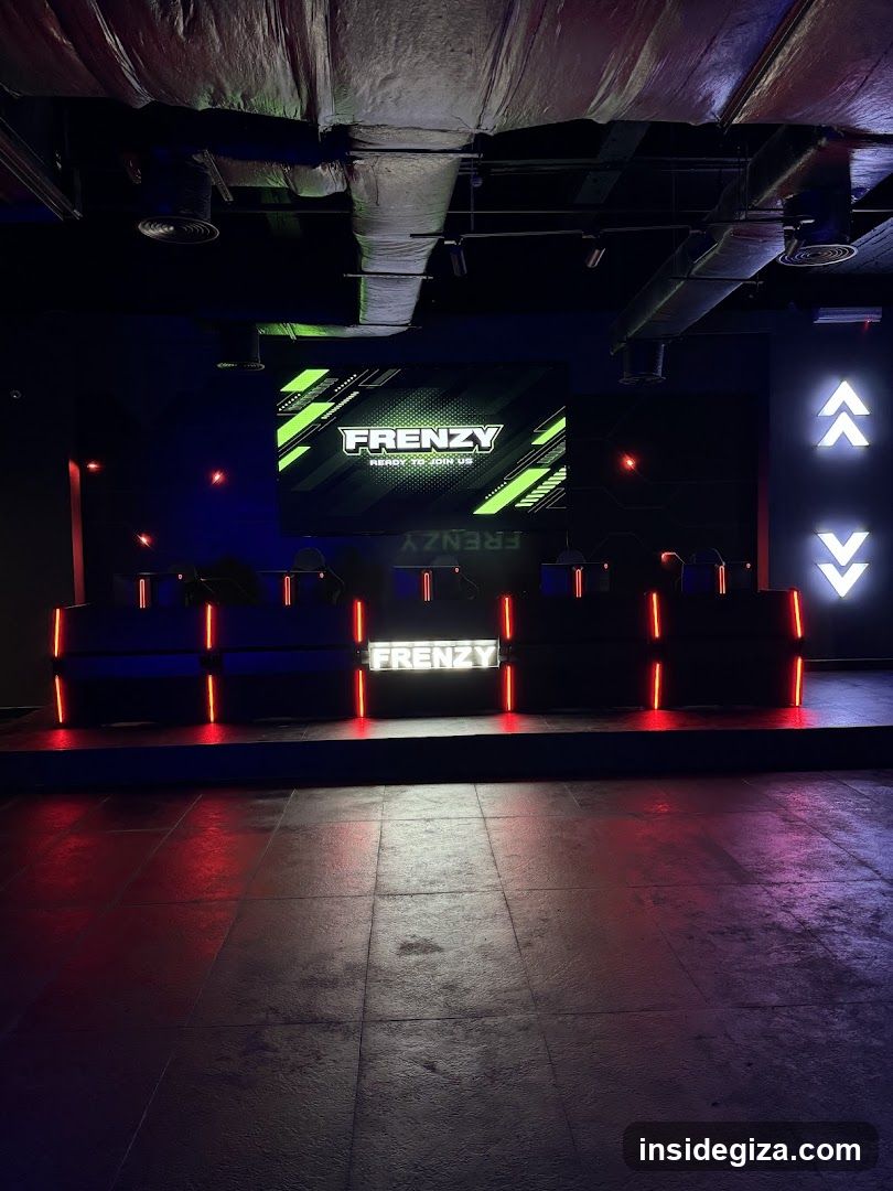 Interior shot of a gaming lounge or arcade with dark ambiance, neon lighting accents in red and green, exposed ceiling pipes, and multiple gaming stations arranged on tiered platforms. A large screen displays the word 'FRENZY' with diagonal stripes. The floor is tiled and reflects ambient light.