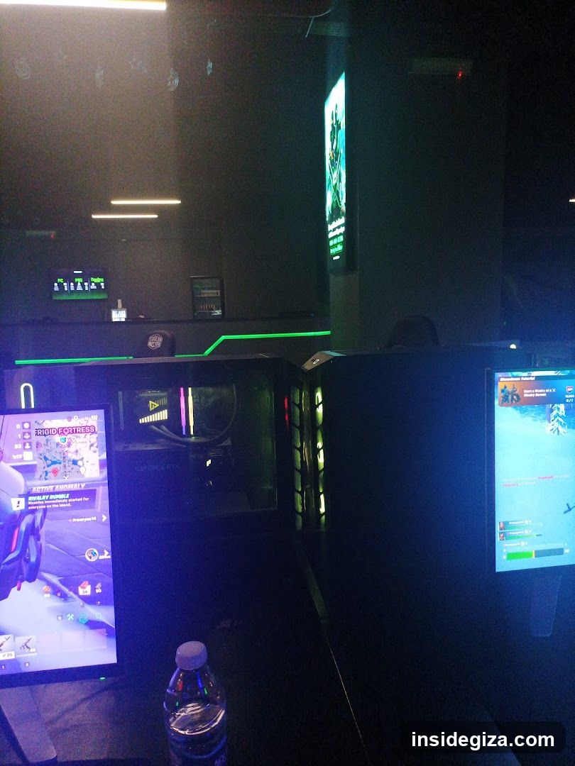 Dark interior of a gaming lounge with multiple monitors displaying games, illuminated by blue and green LED lighting. A water bottle sits on the desk in foreground. Background shows digital signage and ambient glow from screens.
