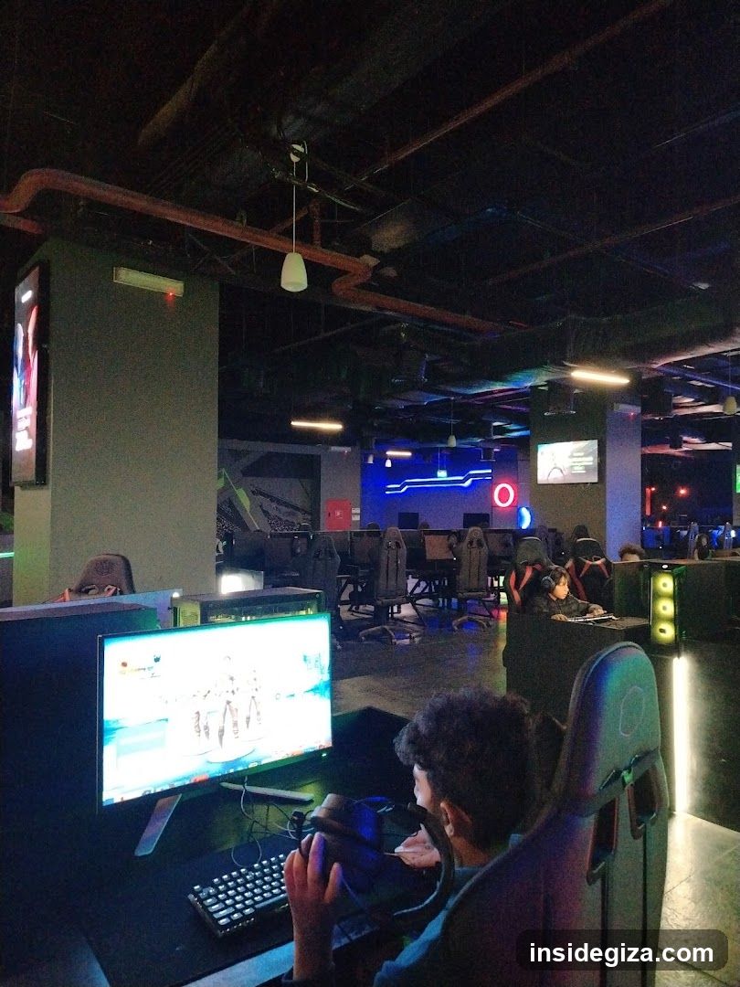 Interior of a gaming lounge with multiple gaming stations, each equipped with monitors and gaming chairs. A person is seated in the foreground playing a game on a monitor displaying a bright screen. The ceiling features exposed pipes and industrial lighting. Neon lights add ambiance to the space.