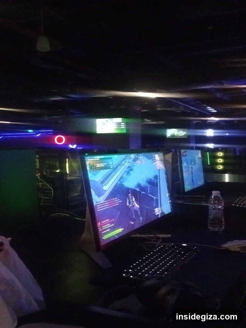 A dimly lit room with multiple computer monitors displaying a video game, likely an FPS or shooter. The setup includes keyboards and water bottles on the desk. Neon lights are visible in the background.