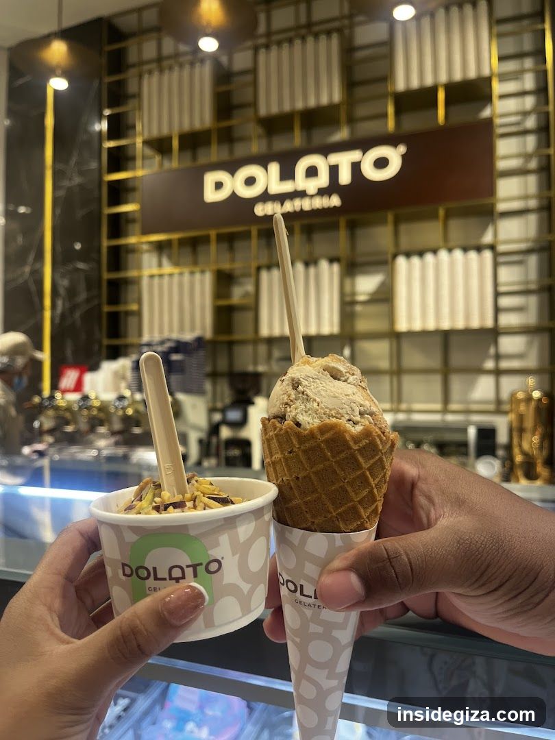 A close-up of two hands holding Dolatto Gelateria products - a cup of ice cream with toppings and an ice cream cone, in front of the shop interior.