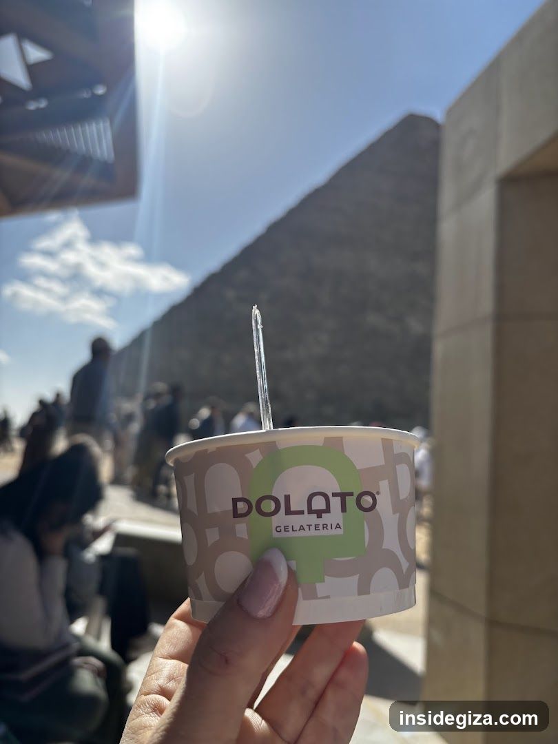 A hand holds a Dolatto Gelateria cup with a straw, set against the backdrop of the Great Pyramid of Giza under bright sunlight.