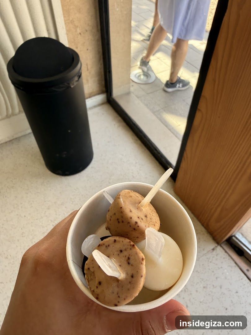 A close-up view of a hand holding a small white paper cup containing two round, speckled ice cream pops and one plain white scoop. The background shows the interior floor with a black trash can and a glass door reflecting the legs of a person standing outside.