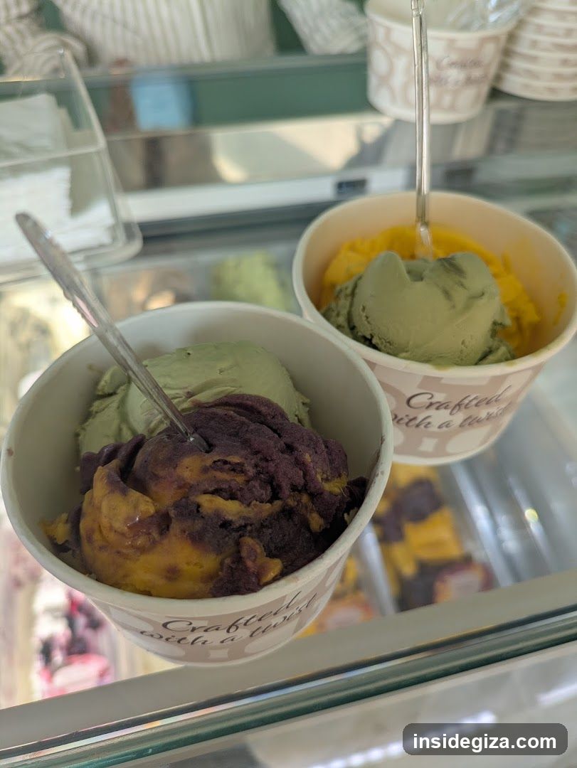 Close-up of two ice cream cups in a glass display case. The cup in the foreground contains a swirl of purple and yellow ice cream, while the one behind it has green and yellow scoops. Both cups have text that reads 'Crafted with a twist'. In the background, more ice cream containers are visible inside the freezer.