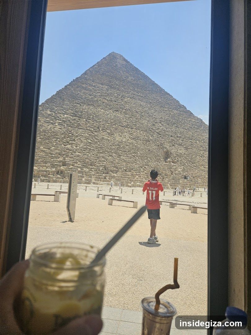 A person holds a jar of food in the foreground, with a view through a window showing the Great Pyramid in the background. A child wearing a red jersey stands near the pyramid.