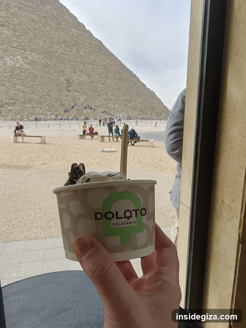 A hand holds a cup of Dolatto Gelateria ice cream in the foreground, featuring visible scoops of chocolate and vanilla flavors. In the background, through a window, is the Great Pyramid of Giza at the Giza Plateau in Cairo, Egypt. The scene captures tourists enjoying gelato near the historic landmark.