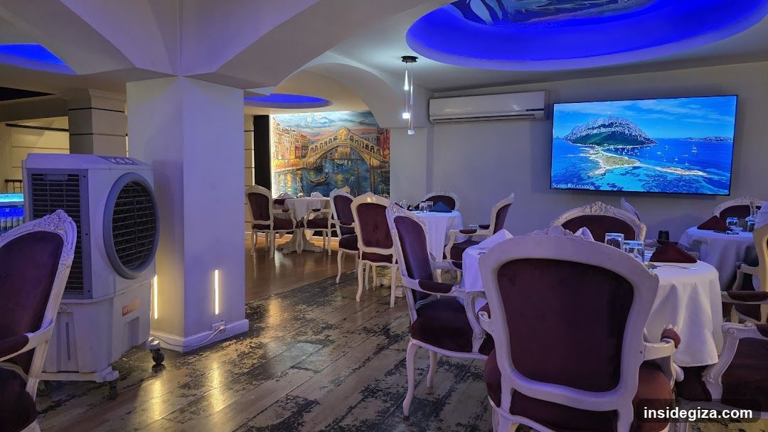 The image shows the interior of a restaurant or dining area, featuring white tablecloths on round tables surrounded by ornate chairs with dark upholstery. The room is illuminated by blue ambient lighting from recessed ceiling fixtures and wall sconces. A large air cooler unit stands near a pillar on the left. In the background, there is a mural depicting a Venetian canal scene. On the right wall, a flat-screen television displays a live feed of a coastal landscape with water and hills. An air conditioning unit is mounted above the screen. The flooring appears to be wood-look tile with a distressed finish.
