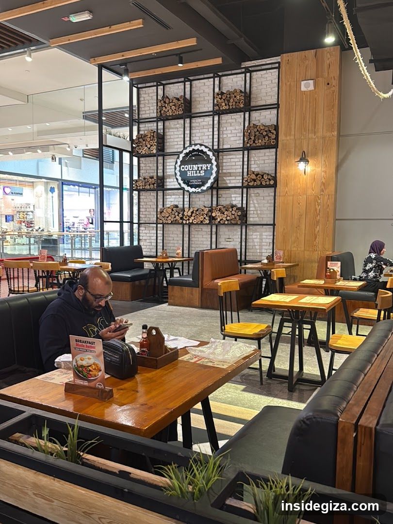 Interior view of a restaurant with wooden tables, black leather booths, and a brick wall decorated with stacked logs. A man sits at a table reading a menu. Visible items include condiments, menus on tables, and decorative plants.