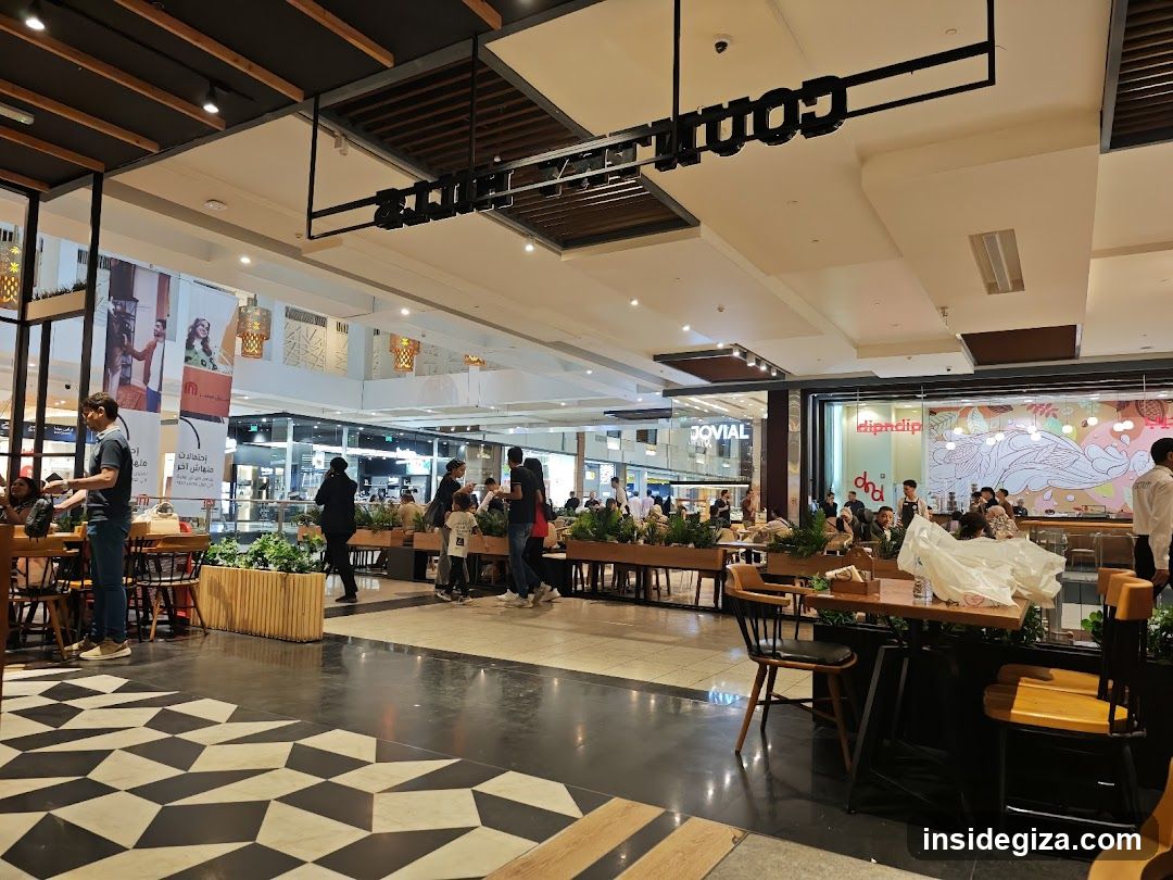 A bustling food court with multiple dining areas, tables, and chairs. Visible elements include a 'COFFEE HUT' sign hanging from the ceiling, various food stalls in the background, and people dining or walking around. The floor features a geometric black and white tile pattern.