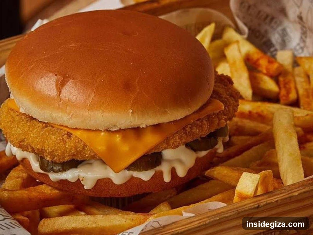 A close-up of a golden-brown cheeseburger served in a wooden basket alongside crispy french fries. The burger features a thick breaded patty, melted yellow cheese, pickles, and creamy sauce on a soft bun.