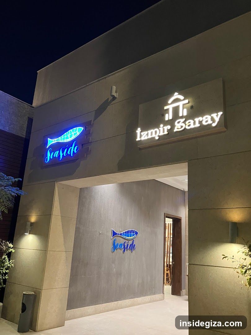 Night photo of a modern building entrance featuring illuminated signage. On the left, a blue neon fish logo with 'Seaside' text below it. To the right, white backlit lettering reads 'Izmir Saray' above an architectural icon resembling a traditional Turkish house. Below, inside the covered entryway, another version of the Seaside fish logo is mounted on the wall. Warm spotlights illuminate the facade and plants flank the sides.