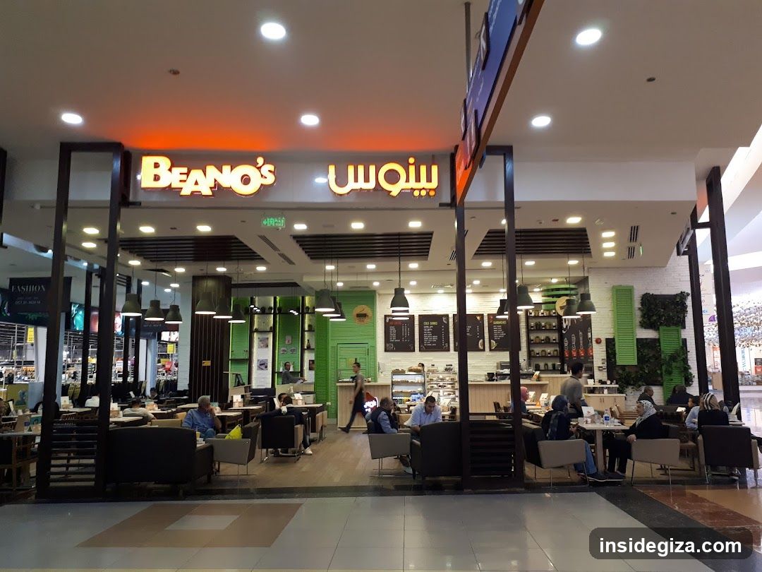 The image shows the exterior of a Beano's coffee shop located inside a shopping mall. The store has a modern design with large glass windows and doors, allowing customers to see into the interior. The signage above the entrance displays 'BEANO'S' in English and Arabic script. Inside, there are tables and chairs arranged for customers, along with a counter area where staff can be seen working. The lighting is bright, creating a welcoming atmosphere.