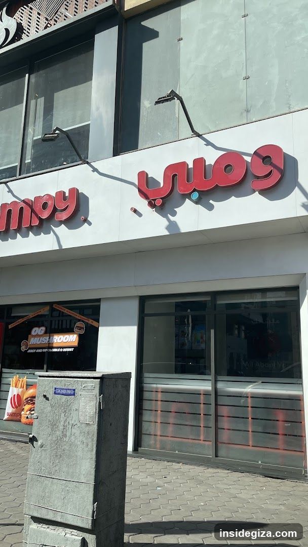 A modern shop front featuring a white facade with red signage displaying 'ومبي' in Arabic script and partial English text ending in 'mpy'. Large glass windows reflect the surroundings, including an orange sign inside reading 'OG MUSHROOM'. A gray utility box stands on the sidewalk in front.