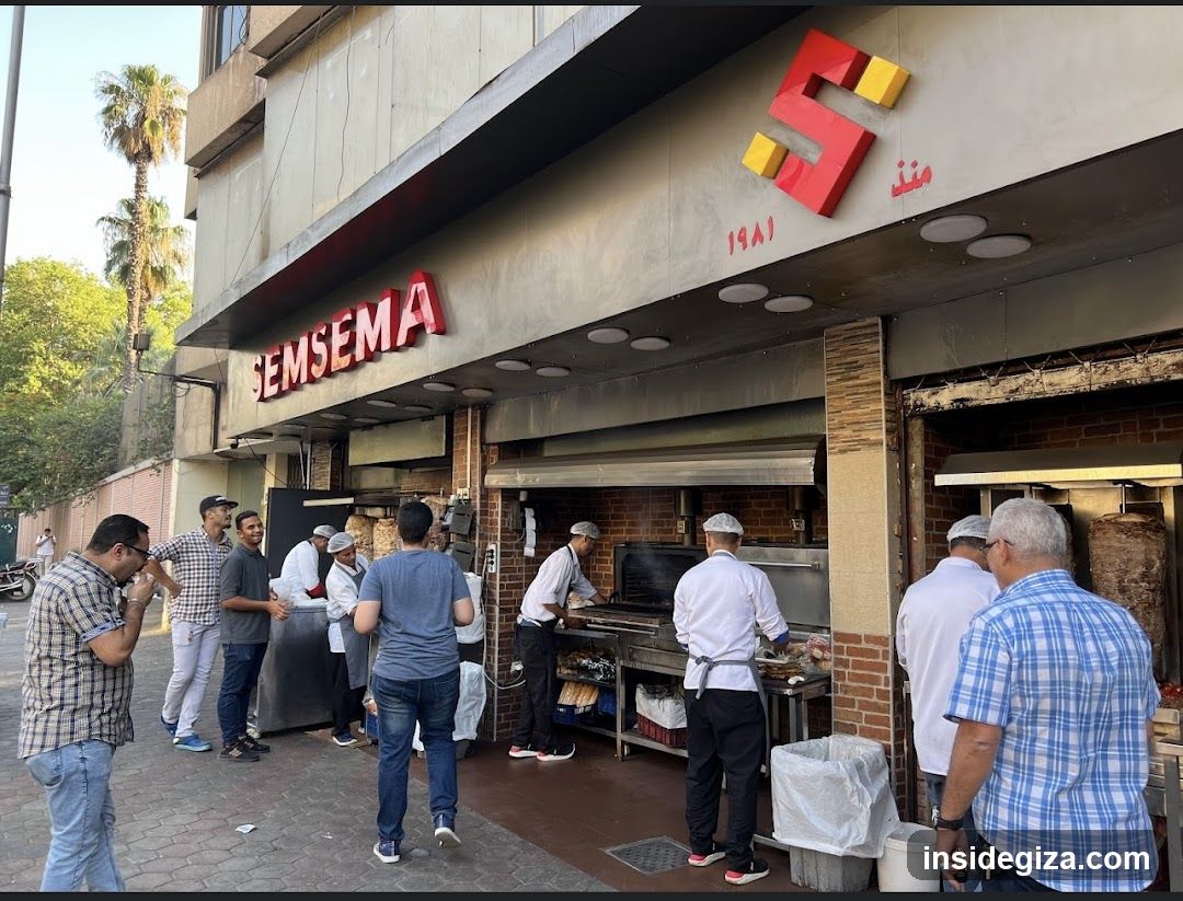 The image shows the exterior of a restaurant named 'SEMSEMA' with Arabic script and the year 1981 displayed on the facade. The building features a modern design with large windows, an open kitchen area where chefs are preparing food, and several people standing outside, some appearing to be customers waiting or ordering. Palm trees in the background suggest a warm climate.