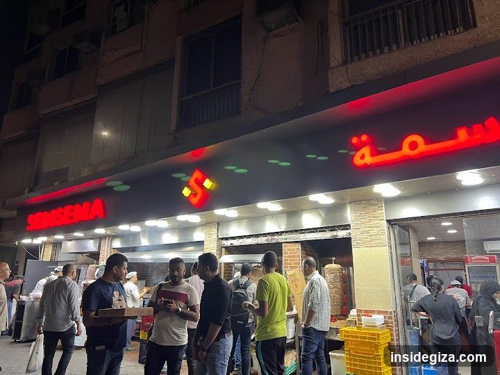 A busy street food stall with illuminated red signage reading 'SESAME' and Arabic script, featuring a queue of customers waiting near the entrance.