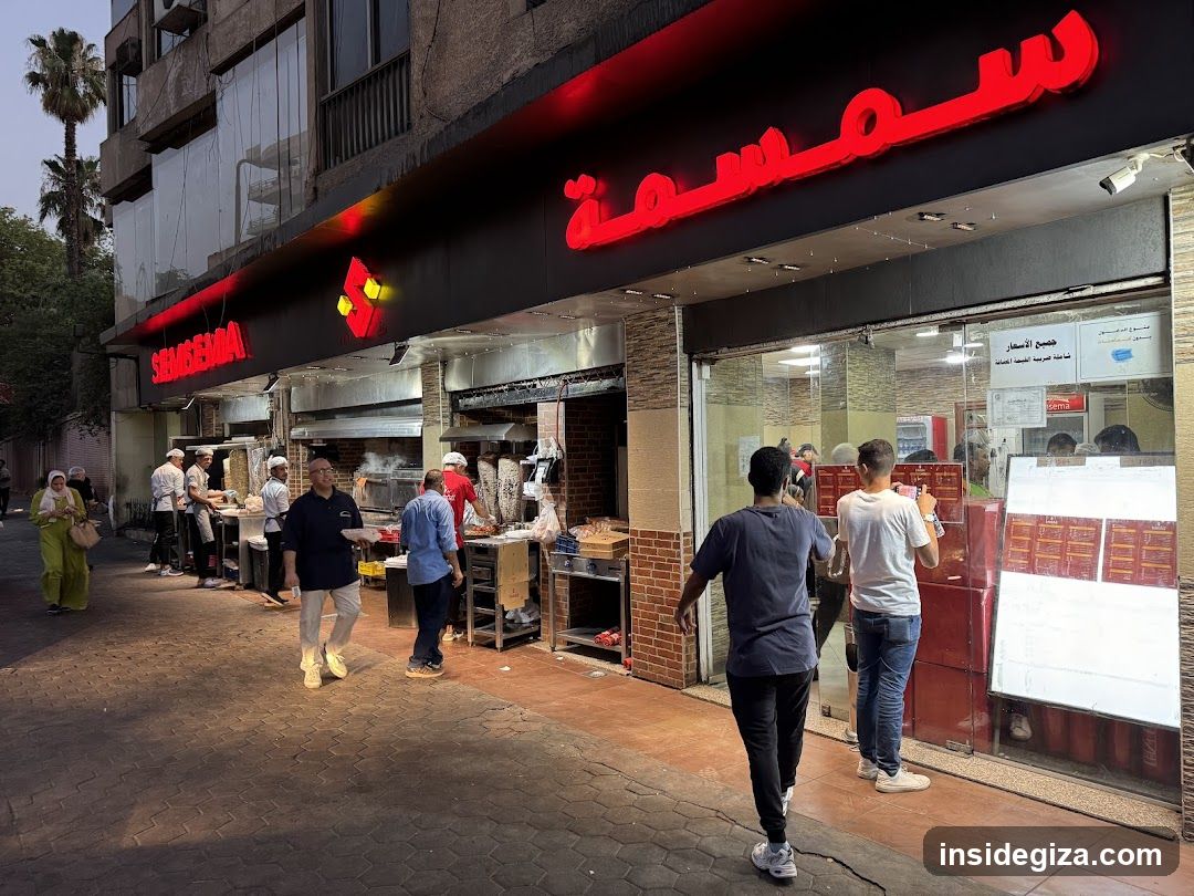 A street-level view of a shop with Arabic signage reading 'سمسمة' (Samosa) in red neon letters above the entrance. The storefront features glass windows and doors, with staff visible inside preparing food. Outside, customers are standing near the counter, some waiting or ordering. A menu board is displayed on the right side of the window.