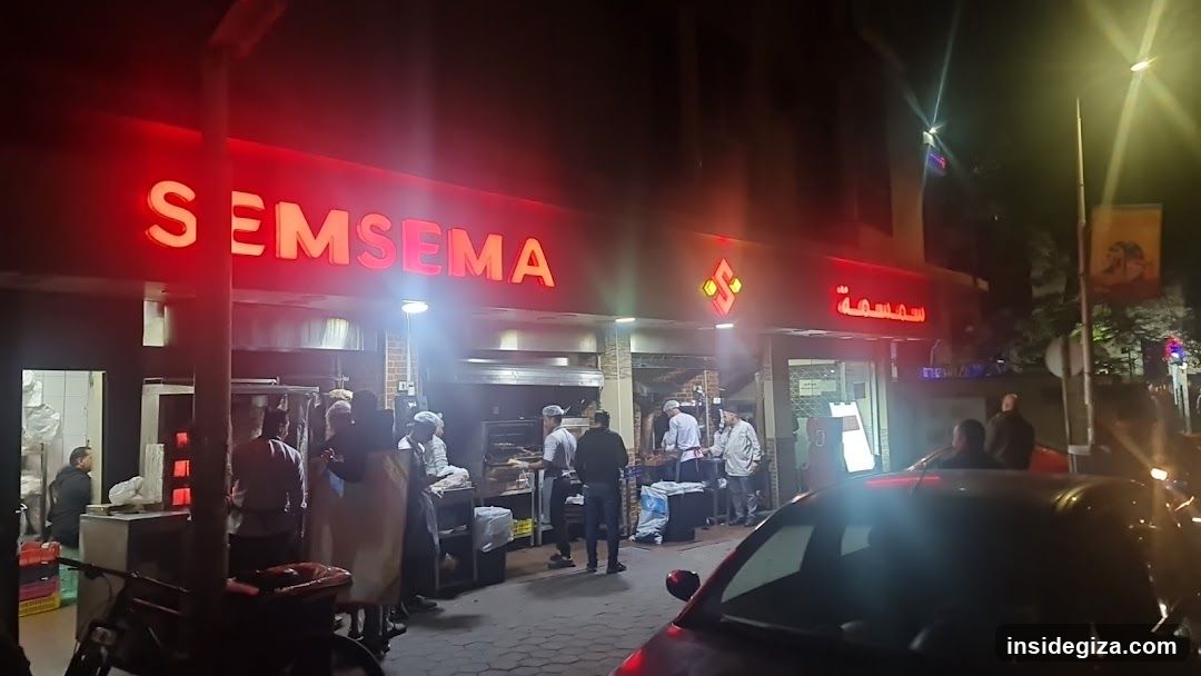 A brightly lit food stall at night with red neon signage reading 'SEMSEMA' and Arabic script below. Several staff members are preparing food behind stainless steel counters, while customers stand nearby waiting or ordering. A car is parked in the foreground, partially obscuring the view. The scene suggests a busy street-side eatery.