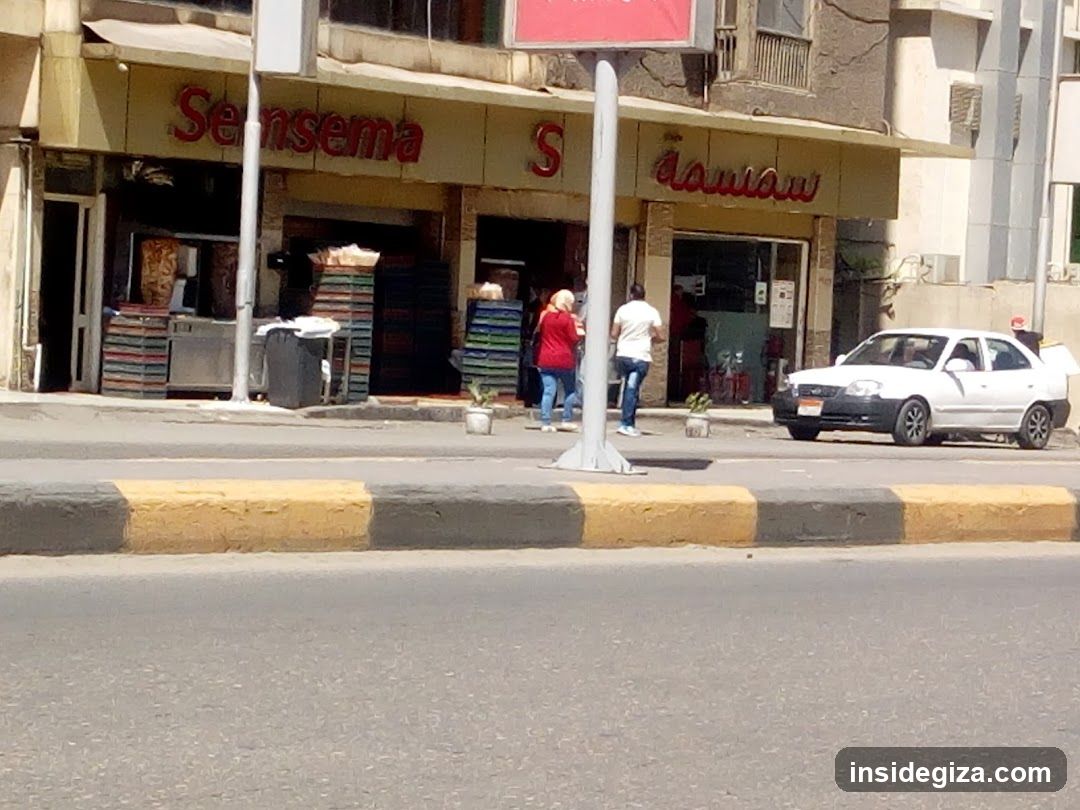 A street-level view of a shop with 'Sensema' signage in red lettering on the facade. The entrance is open, revealing stacked items and people inside. A white car is parked nearby.