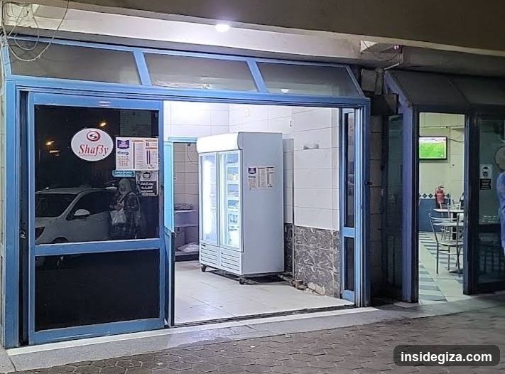 A storefront with blue-framed glass doors and windows, displaying a 'Shafiy' logo on the left window. Inside, there is a white refrigerator unit with illuminated shelves. The interior walls are tiled in white and gray stone. A person wearing a headscarf stands near the entrance. Outside, part of a white car is visible through the glass.