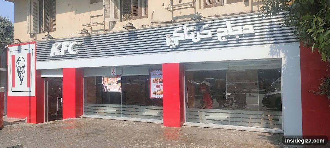Photo of KFC shop front with visible signage including 'KFC' logo and Arabic text on facade, red pillars, glass windows displaying interior seating area, promotional posters inside window showing food items and prices like 99, no menu board or list visible outside.