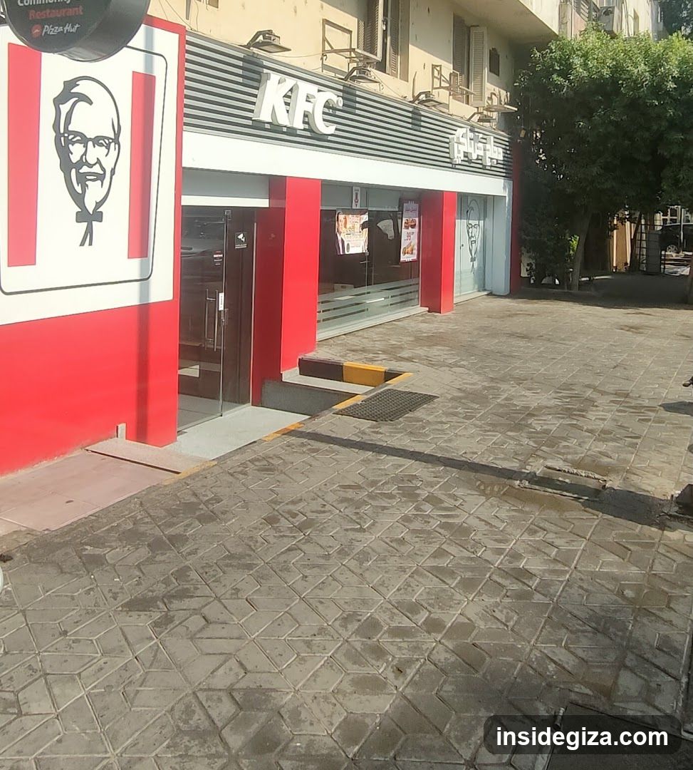 Photo of KFC physical exterior entrance with visible signage on building facade, red and white color scheme, glass doors, and paved sidewalk area.