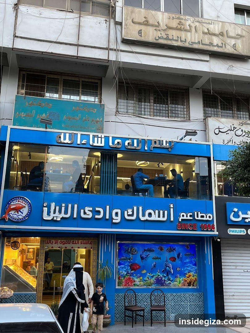 A blue storefront with Arabic signage reading 'مطعم أسماك وادي النيل' (Wadi Al-Nil Fish Restaurant) and 'SINCE 1986'. The facade features a large aquarium mural, two black chairs outside, and people entering or standing near the entrance. A woman in traditional attire walks past the shop.