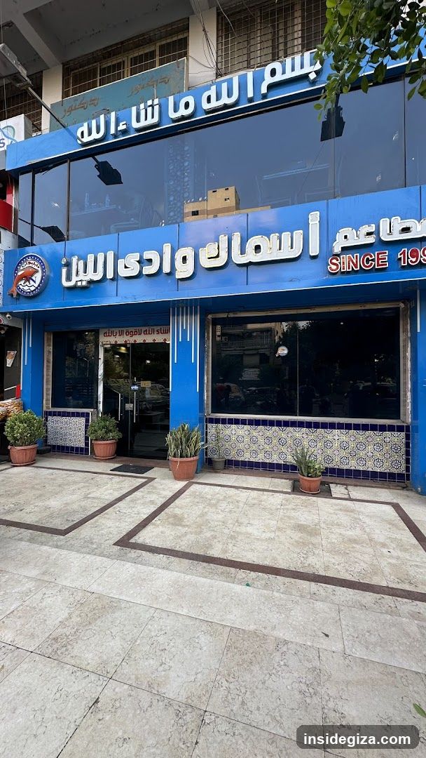 A blue storefront with Arabic signage reading 'Bismillah wa shahada' and 'Asmaak wadi al-bayn', featuring potted plants, tiled base, and glass entrance.
