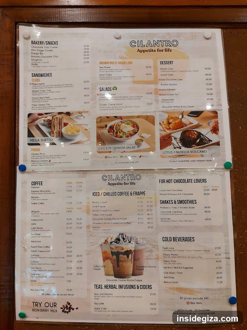 A laminated menu board mounted on a wooden wall, featuring sections like Bakery/Snacks, Sandwiches, Salads, Dessert, Coffee, Iced Drinks, Hot Chocolate Lovers, Shakes & Smoothies, Cold Beverages, and Herbal Infusions.
