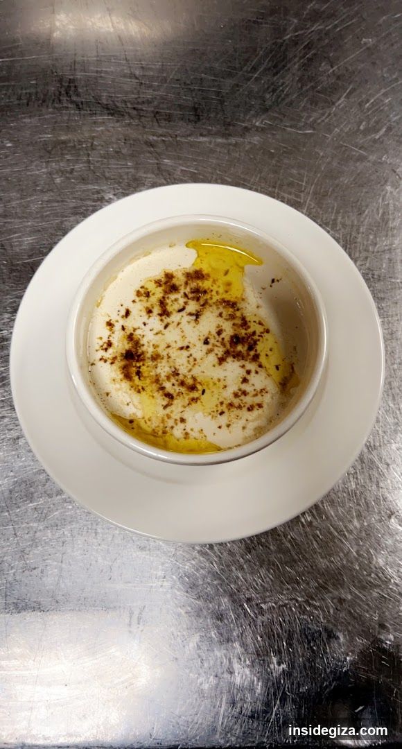 A close-up of a small white bowl containing a creamy, light-colored substance, possibly yogurt or cheese, drizzled with golden olive oil and sprinkled with dark brown spices, likely cinnamon or nutmeg. The bowl rests on a matching saucer atop a scratched stainless steel surface.