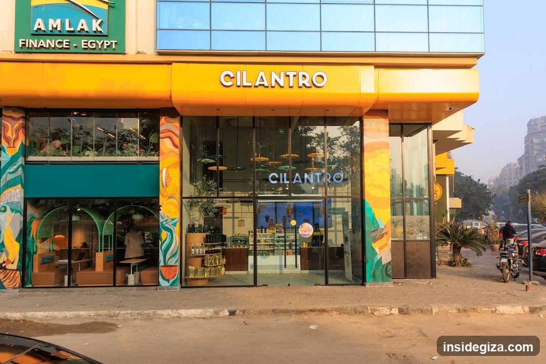 A vibrant yellow storefront with 'CILANTRO' signage above the entrance and on glass doors. The building features colorful mural columns, large windows showing interior seating areas, and adjacent signage for 'AMLAK FINANCE - EGYPT'. A motorcycle is parked nearby on a dusty street.