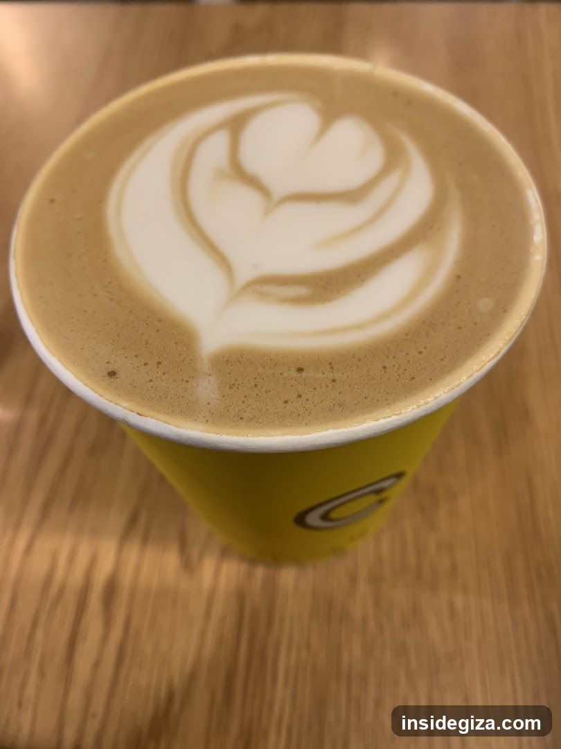 A close-up of a latte with heart-shaped foam art in a yellow disposable cup, placed on a wooden table surface.