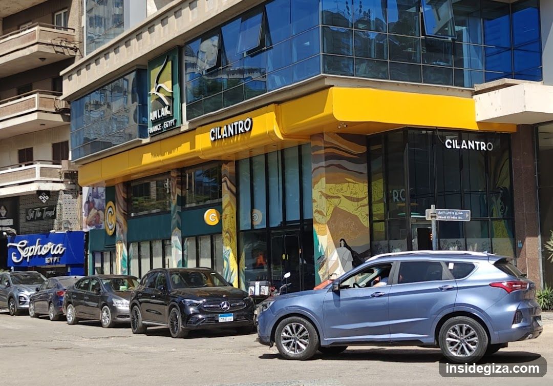 A modern shop front featuring a bright yellow awning and signage reading 'CILANTRO' on the building facade. The entrance is flanked by glass doors and windows, adorned with colorful murals depicting abstract designs and figures. A blue SUV is parked in front of the store, alongside other vehicles along the street. Adjacent to the Cilantro store is another establishment named 'Sephora', visible through its blue sign.