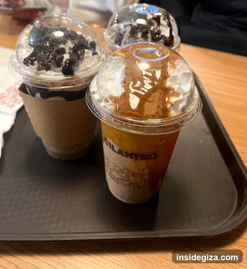 Three iced beverages in clear plastic cups with dome lids, placed on a black tray atop a wooden table. The cup in the foreground features 'CILANTRO' branding and caramel drizzle over ice; others contain toppings like chocolate chips or pudding.