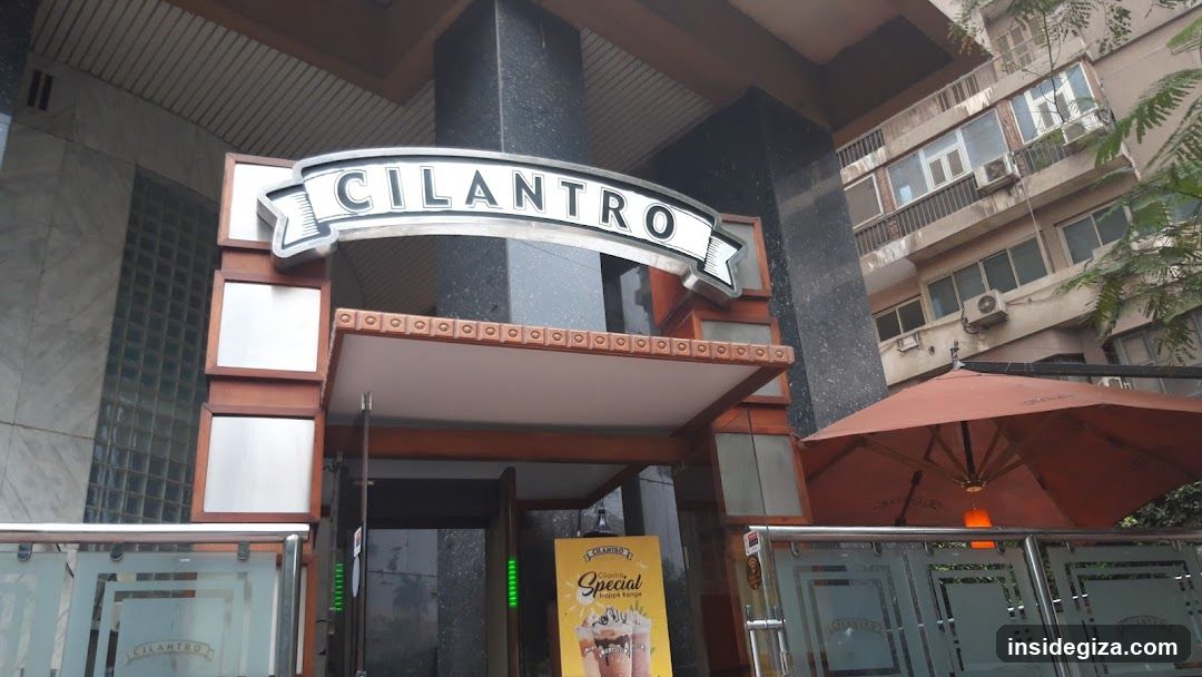 Exterior view of Cilantro restaurant entrance featuring a prominent arched sign with the name 'CILANTRO' mounted above the doorway. The facade includes dark marble columns, wooden trim accents, and glass panels on either side. A promotional poster for a special drink is visible near the entrance. An orange umbrella shades part of the outdoor seating area to the right.