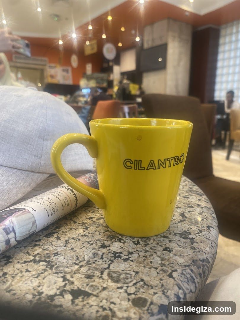 A yellow mug with 'CILANTRO' printed in black letters sits on a speckled granite table next to a rolled-up newspaper or magazine. In the background, there is an indoor setting with warm lighting, hanging bulbs, and people seated at tables.