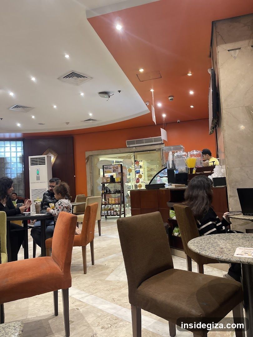 Interior of a restaurant or café with orange and white ceiling accents, marble flooring, and patrons seated at tables. A staff member is visible near the counter area in the background.