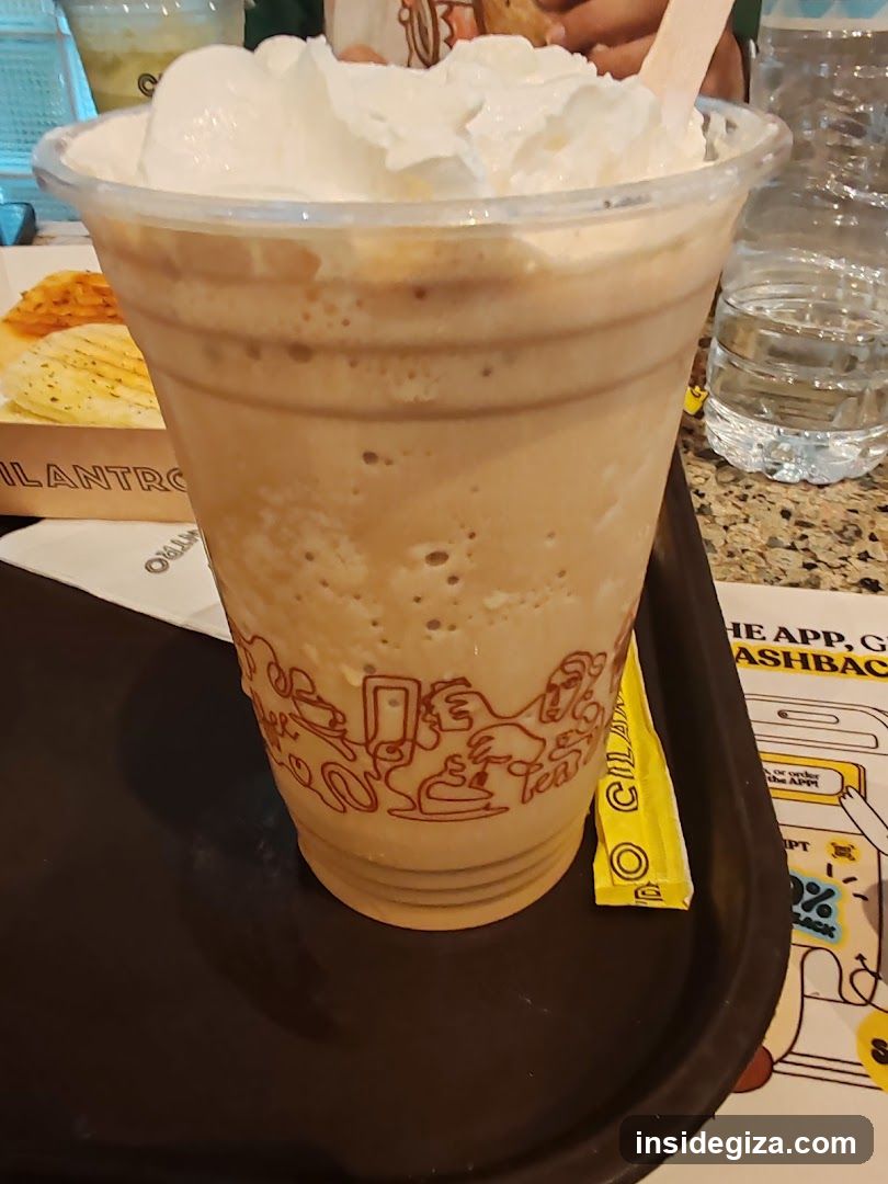 A close-up of a tall plastic cup filled with an iced coffee beverage topped with whipped cream and a wooden stirrer. The cup has red line art branding on it, including cartoon figures and text that appears to say 'O'Kee'. It sits on a black tray next to a yellow packet (possibly sugar or syrup) and a printed flyer or menu insert in the background showing partial text like 'THE APP', 'CASHBACK', and '10% OFF'. A water bottle is visible in the background.