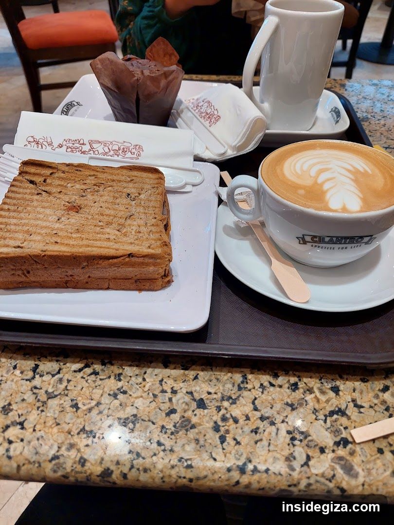 A tray with a latte art coffee, a grilled sandwich, a pastry in a brown paper wrapper, and a white mug on a granite countertop.