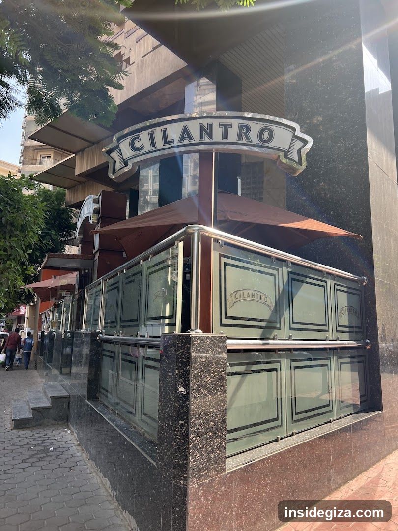 The image shows the exterior of a restaurant named 'CILANTRO', featuring a prominent sign above the entrance. The building has a modern design with glass panels and metal railings surrounding an outdoor seating area. Brown umbrellas provide shade over the tables, and pedestrians can be seen walking on the sidewalk nearby.
