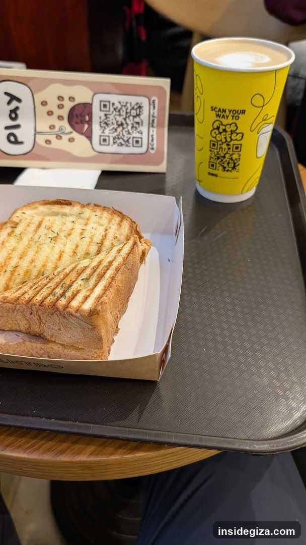 A tray with a grilled sandwich in a paper container, a yellow cup of coffee with latte art, and a QR code card labeled 'play'.